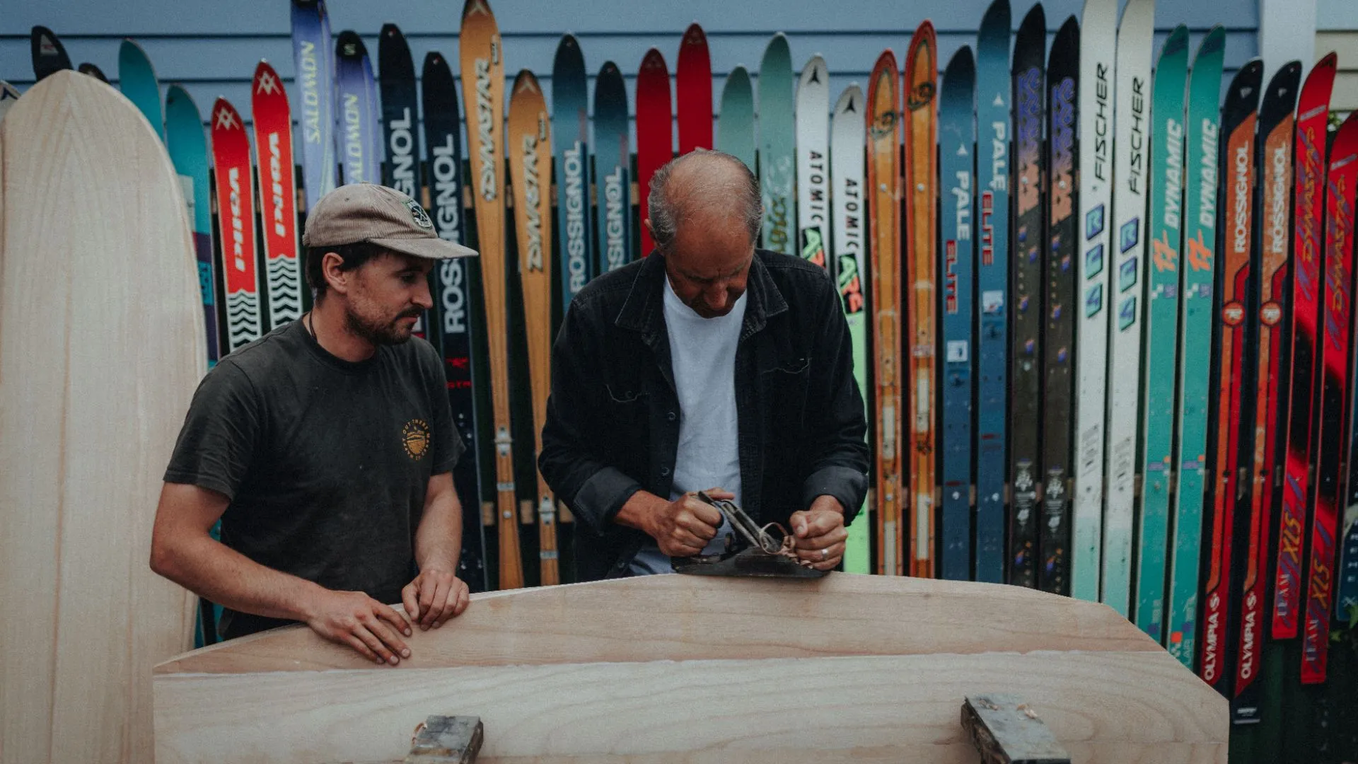 Two builders planing a timber blank in front of a fence of old skis